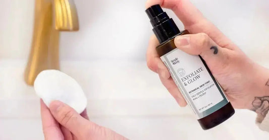 A hand holding a bottle of Simple Body Exfoliate and Glow Botanical Skin Tonic misting it onto a cotton pad with a gold faucet in the background.