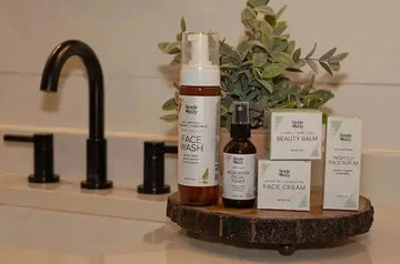 A photo of Simple Body products sitting on a wooden tray including Chamomile Foaming Face Wash, Rosewater Facial Toner, Beauty Balm, Face Cream and Nightly Face Serum with a green plant in the background and a black modern faucet.