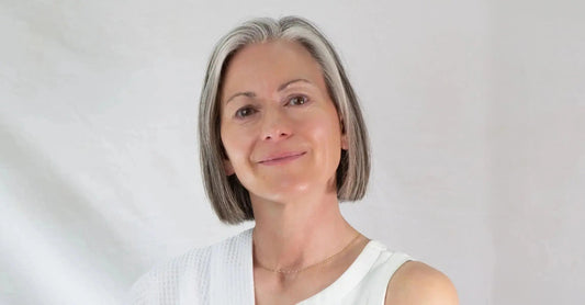 A woman in her late fifties with glowing skin and gray hair smiling at the camera.