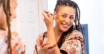 A young woman looking in the mirror smiling with beautiful dreadlocks applying a Simple Body Caramel Clay Mask.