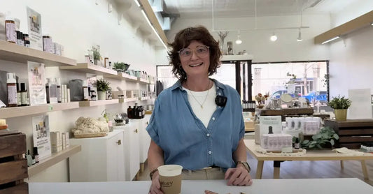 A photo of Jewels Burdick, Founder of Simple Body, standing in the retail store holding a cup of coffee and smiling.