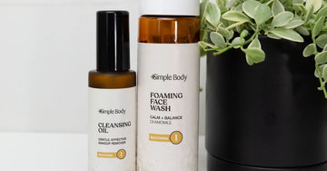 A close up image of Simple Body Cleansing Oil and Simple Body Chamomile Foaming Face Wash sitting next to a black ceramic with a black pot and a vibrant green plant