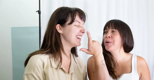 Two young ladies laughing and having fun with one of the girls applying Simple Body Face Cream on the others nose while she scrunches her face.