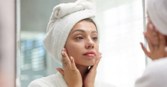 A woman looking in the mirror looking calm and holding the sides of her face with her hair wrapped in a white towel.