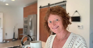 A photo of Jewels, Simple Body Founder standing in a kitchen with a cup of coffee wearing a white crochet sweater.