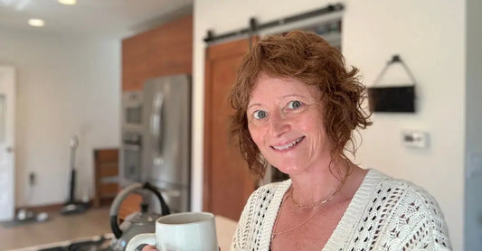 A photo of Jewels, Simple Body Founder standing in a kitchen with a cup of coffee wearing a white crochet sweater.