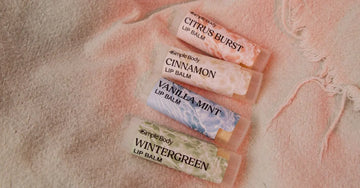A photo of Simple Body Lip Balms including Citrus Burst, Cinnamon, Vanilla Mint, and Wintergreen laying on a beige cloth.
