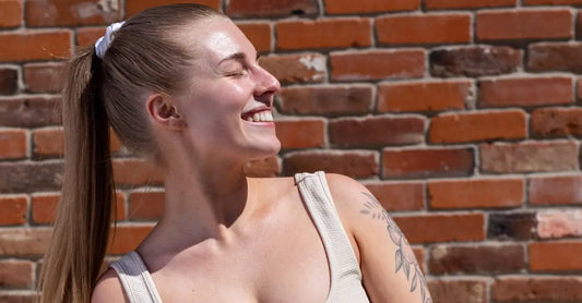 A photo of Caitlyn looking into the sunshine and smiling. She wears a ponytail for her long hair and an off white tank top and sits in front of a brick wall.
