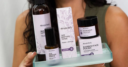 A photo of a woman holding a green glass tray with Simple Body products balanced on it including a Lavender Body Lotion, Age Defense Serum, Age Defying Tonic, Lavender Sugar Scrub, and Bamboo Face Polish.