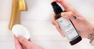 A hand holding a bottle of Simple Body Exfoliate and Glow Botanical Skin Tonic misting it onto a cotton pad with a gold faucet in the background.