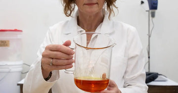A close up image of Jewels Burdick, Founder of Simple Body in her lab coat holding a glass beaker looking at an orange colored oil with her lab in the background.
