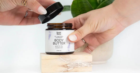 A pair of hands with white nail polish removing the lid from a Simple Body Lavender Body Butter sitting on a natural wooden block with a plant in the background.
