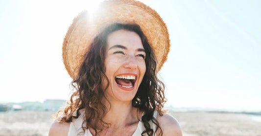 A beautiful woman laughing standing in front of the ocean on a bright summer day wearing a straw hat.