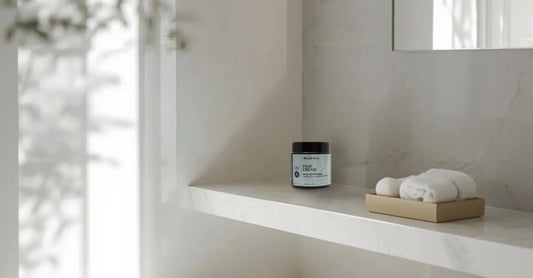 A minimalist photo of a white bathroom with a clean, white shelf, a bamboo towel holder with two white towels and a jar of Simple Body Face Cream in the corner.