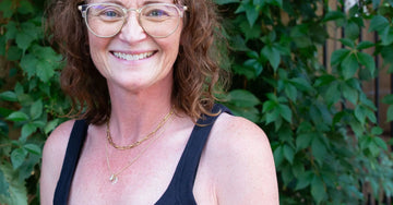 A photo of our founder, Jewels, looking into the camera and smiling with gold framed glasses and a black dress.