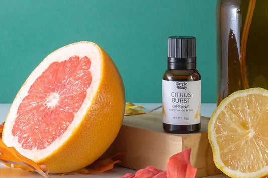 A photo of a bottle of Simple Body Citrus Burst Essential Oil surrounded by a half grapefuit, half orange and an amber colored bottle and fall leaves with a green background