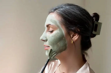A photo of a young woman with dark hair pulled back in a hair clip applying a Simple Body Green Clay Mask with a Fan Brush and her eyes closed on a neutral background