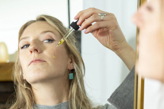 A photo of a young woman looking in the mirror applying Simple Body Pomegranate Oil to her cheek with beautiful skin.