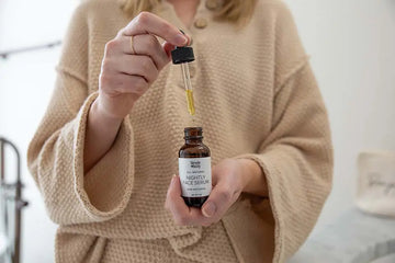 A photo of a woman in a beige lounge outfit holding a bottle of Simple Body Nightly Face Serum with the dropper pulled out so you can see the golden oil. 