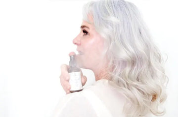 A photo of Erin, a mature woman with beautiful gray hair looking to the side with radiant skin misting a Simple Body Rosewater Toner over her shoulder.