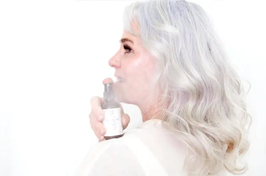 A photo of Erin, a mature woman with beautiful gray hair looking to the side with radiant skin misting a Simple Body Rosewater Toner over her shoulder.