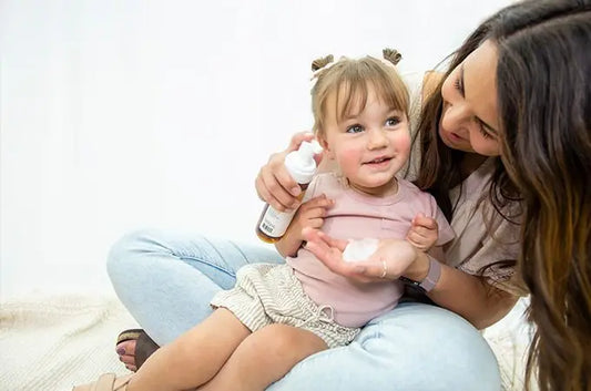 A photo of a mom with long, dark hair holding her toddler and a bottle of Simple Mama Gentle Foaming Shampoo and Body Wash with foam in the other hand. Both are smiling.
