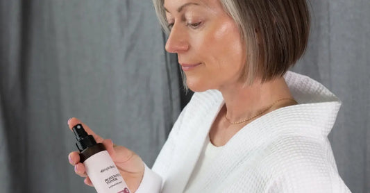 A photo of a mature woman holding a bottle of Simple Body Age Defying Botanical Skin Tonic with glowy, beautiful skin and gray hair.