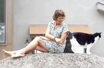 A photo of Jewels Burdick, Founder of Simple Body, sitting on a large rock playing with her tuxedo cat, Alex.