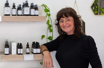 A photo of the Founder of Simple Body, Jewels leaning on a shelf that holds the new Botanical Skincare Tonics and High Performance Serums
