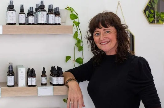 A photo of the Founder of Simple Body, Jewels leaning on a shelf that holds the new Botanical Skincare Tonics and High Performance Serums