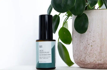 A photo of Simple Body Age Defense High Performance Serum sitting next to a beige ceramic pot holding a green plant.