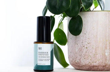 A bottle of Simple Body Barrier Defense Serum sitting next to a beige ceramic pot with a green plant in it.