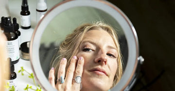 A photo of a young lady looking in the mirror feeling her soft skin from using her products in the background which are Simple Body Nightly Face Seru, Simple Body Pomegranate Oil and Simple Body Foaming face Wash.