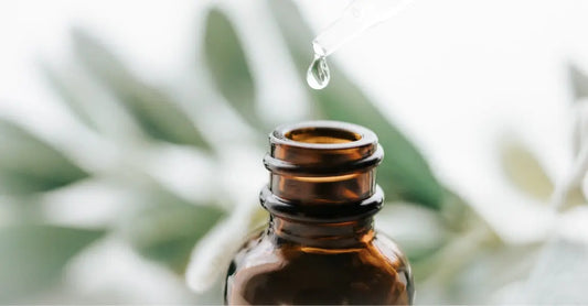 A photo of an amber colored bottle with a blurry plant in the background and a drop of clear serum dropping into the bottle