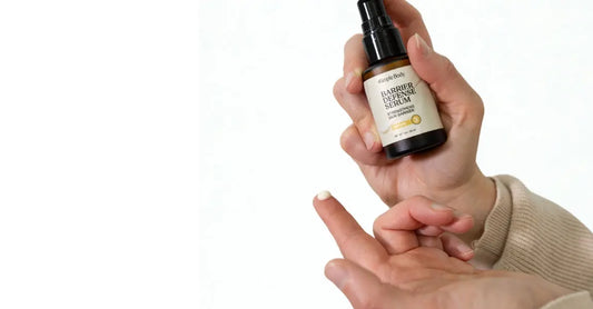 A photo of a hand holding a bottle of Simple Body Barrier Defense Serum. There's a drop of the Barrier Defense Serum on her fingertip showing off the creamy white texture.