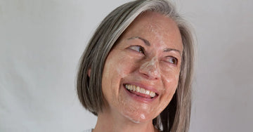 An upclose photo of a woman in her mid 50's with a gray bob smiling and looking right with foaming cleanser on her face.