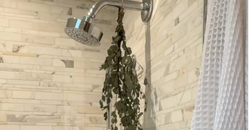 A photo of a shower with marble tile surround, a chrome rainhead shower, dried eucalyptus hanging from the showerhead and a white waffle weave towel in front.