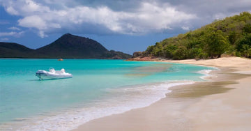 A photo of the beautiful blue teal waters of Antigua in the Carribean with a lone boat, sandy beach and green trees with mountains in the background.