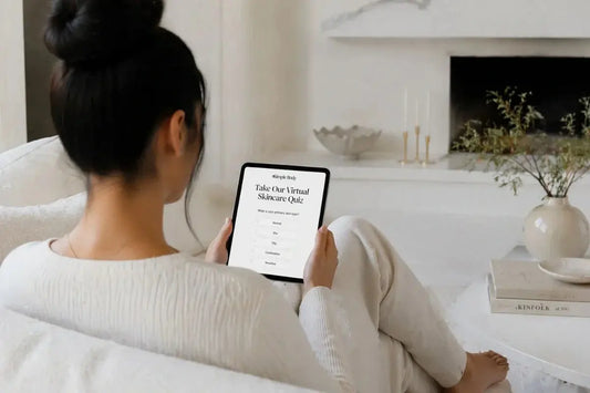 A woman sitting on a white sofa taking the Simple Body Skincare Quiz with a white fireplace, candles and a ceramic vase with a green plant.