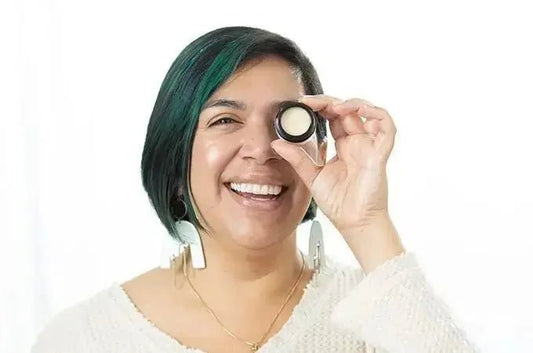 A photo of Ani, a customer of Simple Body smiling with teal streaks in her hair holding a bottle of Simple Body Face Cream and smiling big.