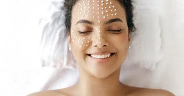 A young woman laying down with her eyes closed, smiling with dots of Simple Body Face Cream on her skin.