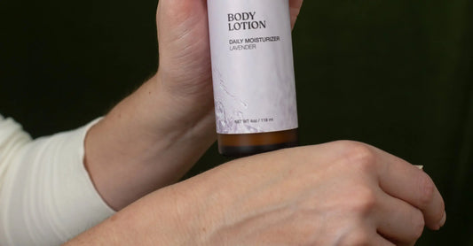 A close up of a hand holding a bottle of Simple Body Lotion in Lavender getting ready to apply to her other hand with a black background.