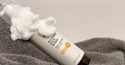 A photo of Simple Body Foaming Face Wash for Sensitive Skin with a yellow Chamomile label laying on a gray towel with dense cleansing foam on top