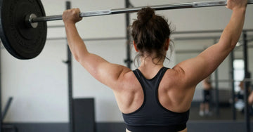 A woman lifting a barbell over her head with a black sports top on that show back acne