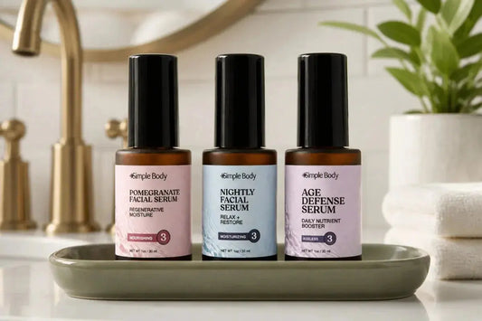 Bottles of Simple Body Pomegranate Facial Serum, Nightly Face Serum, and Age Defense Serum in a green tray on a bathroom countertop and gold faucet in the background.