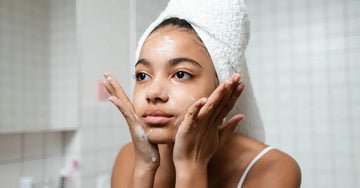 A young woman looks in the mirror with a towel wrapped around her head applying a Simple Body Foaming Face Wash and appearing to massage her skin