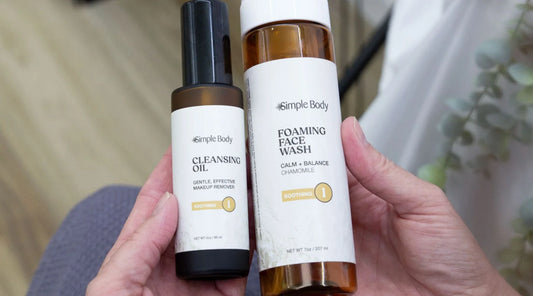 A close up image of hands holding a bottle of Simple Body Cleansing Oil and Simple Body Foaming Face Wash in Chamomile with a blurred background.