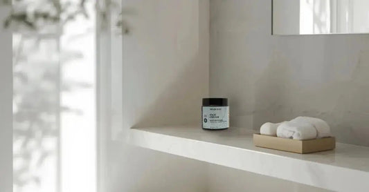 A minimalist photo of a white bathroom with a clean, white shelf, a bamboo towel holder with two white towels and a jar of Simple Body Face Cream in the corner.