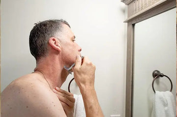 A photo of a man looking in the mirror shaving with a single blade safety razor