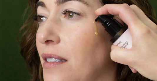 A woman looks to the side as Simple Body Pomegranate Facial Serum is applied to her cheek to her dry skin with a green background.